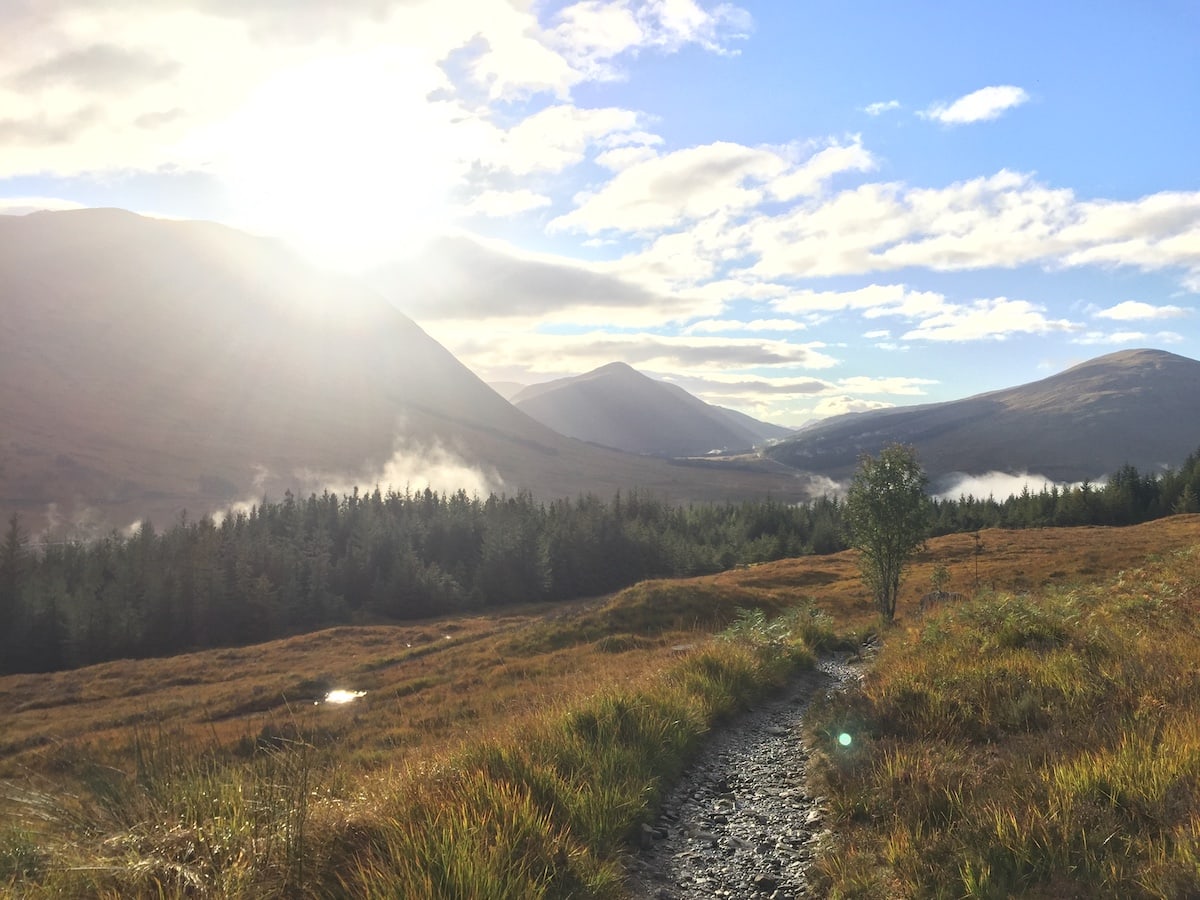 The sun rises over a valley, a forest and a rocky trail in the scottish highlands.