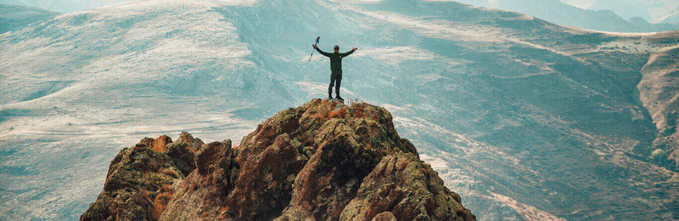 A hiker celebrating reaching the summit after a climb with rolling hills in the background.