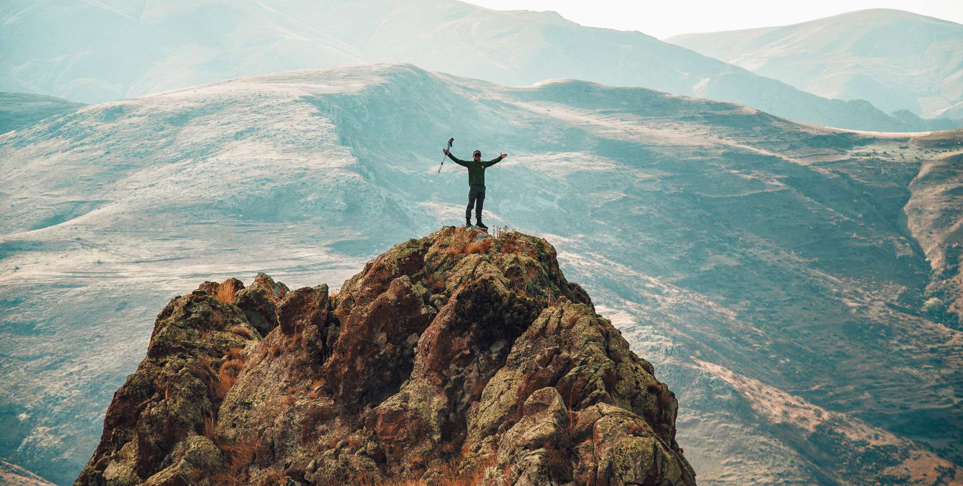 A hiker celebrating reaching the summit after a climb with rolling hills in the background.