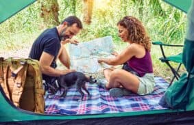 Two people sitting outside a tent holding a map with a dog by their side.