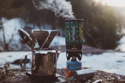 A Jetboil camping stove and a metal coffee pot on the ground with snowy nature in the background.