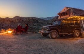 Black jeep camper with fairy lights and a couple seated by a fire in the desert.