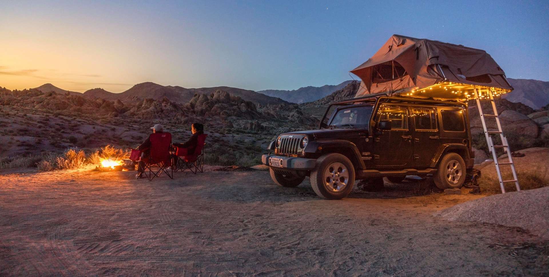 Black jeep camper with fairy lights and a couple seated by a fire in the desert.