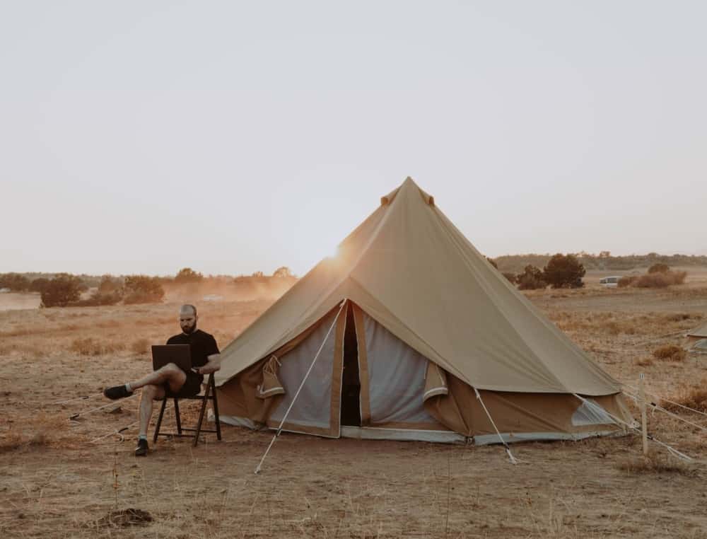 A backpacker on a laptop sitting beside a canvas tent using the tent's shade.