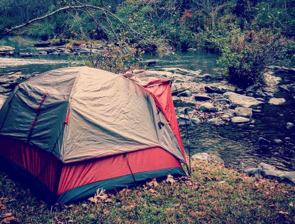 A red and gray tent set up by a river in the fall