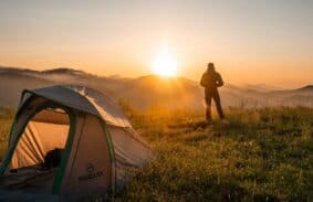 A man stands as a silhouette against a sunrise with a tent in the foreground.