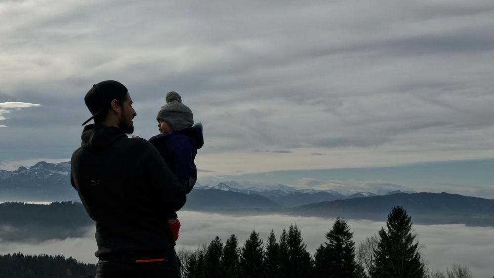 Man holding a baby wrapped up warm on a ridge overlooking mountain in the clouds
