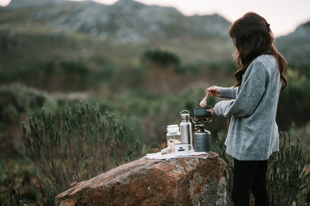 Woman cooking a meal using a small camp stove on a rock in a mountain meadow.