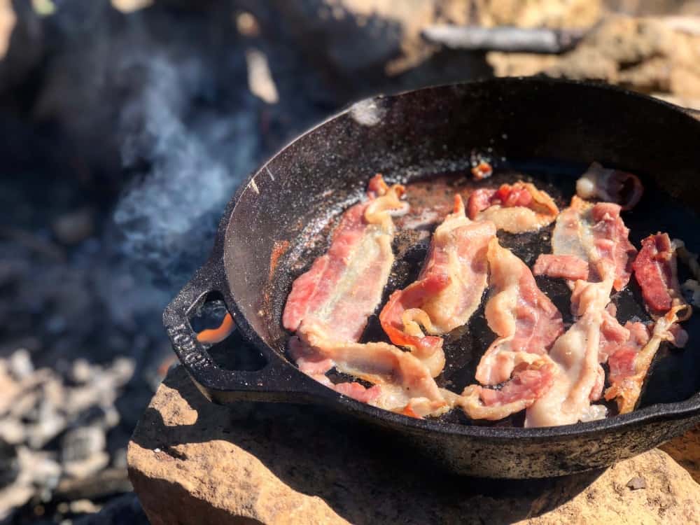 Bacon cooking in a cast iron camping pan on a campfire.