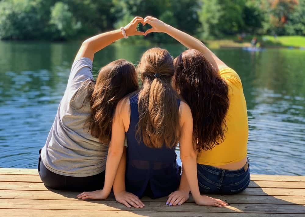 Three women sitting on a dock on a lake with the outer two making a heart with their hands above the middle woman.