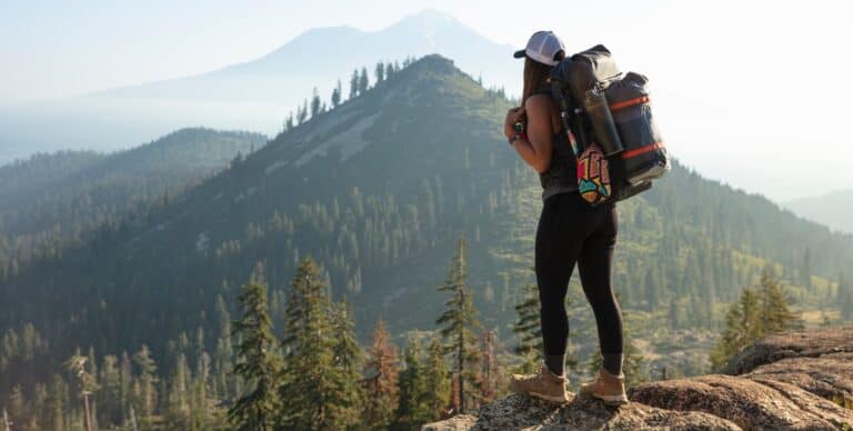 Woman backpacking with a full backpack admiring the view from a hill across to more forested hills in the distance.