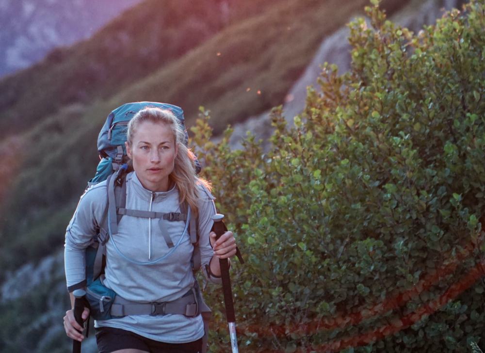 A woman carrying a backpack hiking up a mountain with trekking poles.