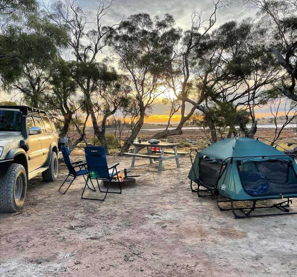 A double camp cot at a beach location with a table and chairs beside a SUV.