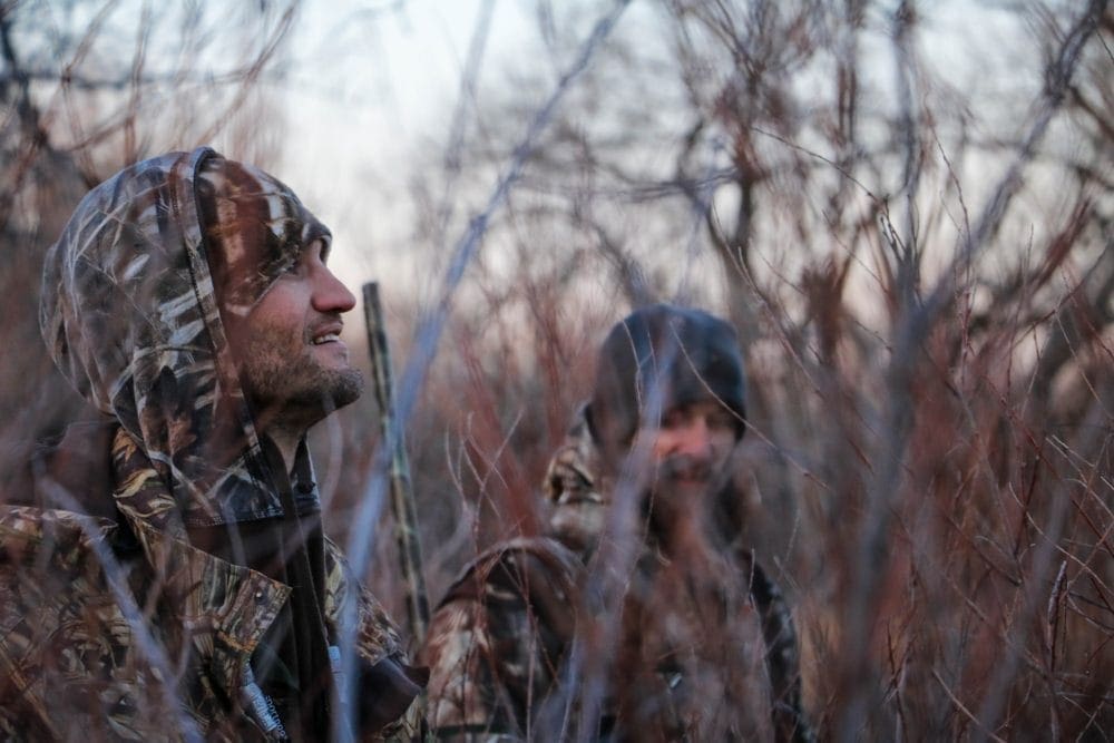 Two hunters in shrubs dressed in camo gear looking for deer