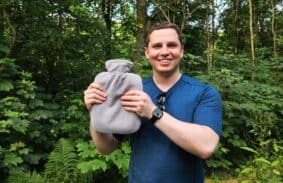 A camper standing in a forest holding a hot water bottle that he is using to keep warm at night.