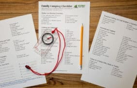 Photo of a printed family camping essentials checklist with compass and pencil lying on a wooden table